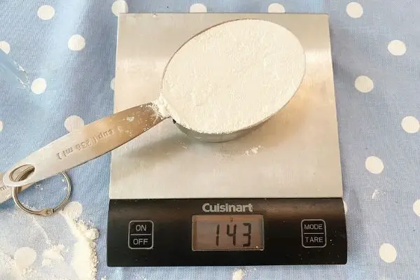 Xicara de farinha na balança pesando 143 g
